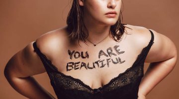 Oversize female in black bra on brown background. Woman with words "You are beautiful" written on her body.