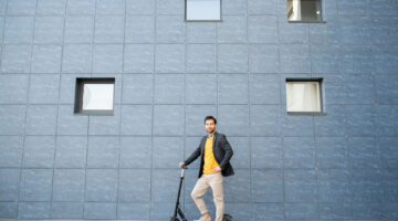 Young well-dressed businessman in smart casualwear standing by wall of modern building while going by scooter