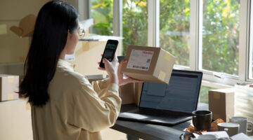 Asian female small business owner using mobile app on smartphone checking parcel box. Warehouse worker, seller holding phone scanning retail dropshipping package postal parcel bar code on cell technology.