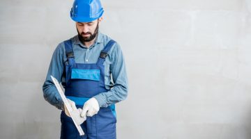 Plasterer in blue working uniform cleaning spatula standing on the grey wall background