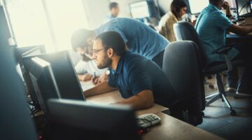 Group of mid 20's and 30's men and women engaged in application development job. They are seated by long desks back to back, each person in front of dual screen computer. Bad posture is innevitable. Back lit, toned image.
