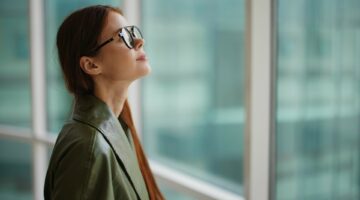 Fashion woman in a stylish raincoat and sunglasses standing by the window and looking at the city and the street. High quality photo