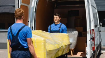 Selective focus of movers holding sofa in stretch wrap near truck on urban street